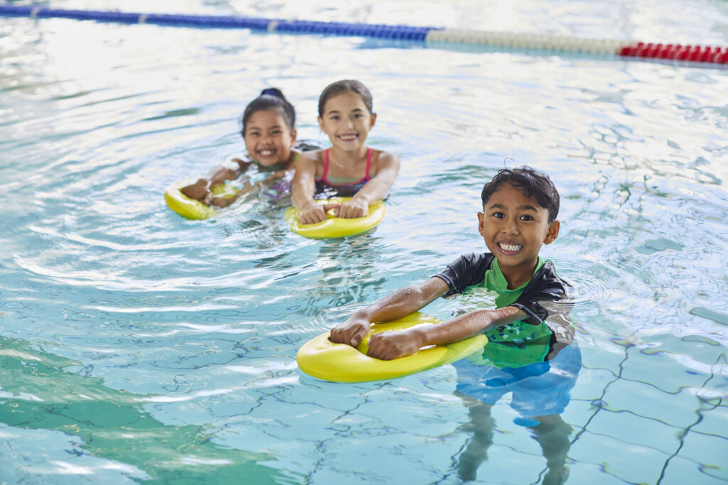 Photo of three young students of diverse gender and background in the swimming pool, each holding yellow kickboard and smiling.
