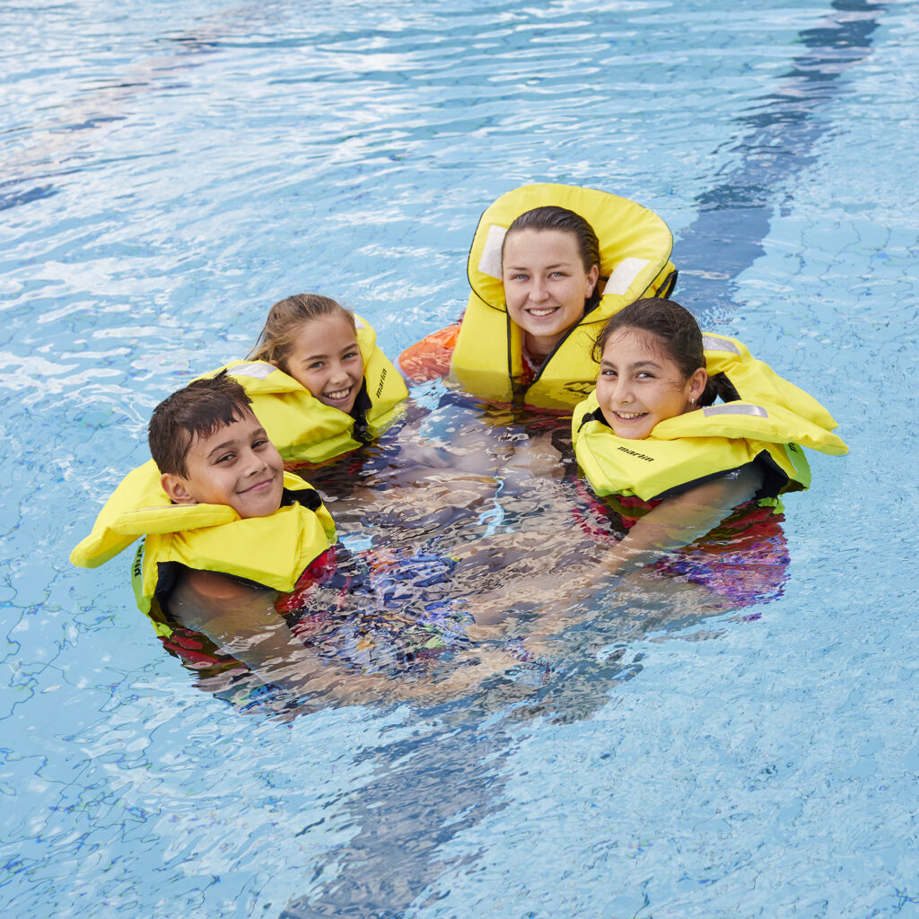 Photo of three young students of mixed gender, linking arms with their swim teacher, all smiling and wearing life jackets in the swimming pool.
